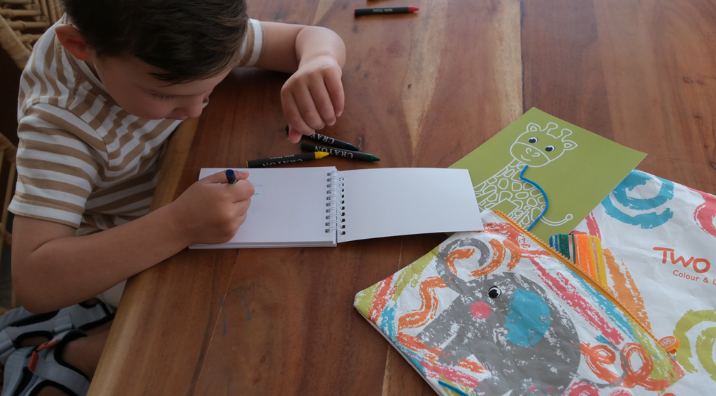 Child drawing in a notebook with colourful crayons and activity cards, featuring TWO CUBS travel essentials like a kids' backpack and Crafty Curls for creative play during school holidays