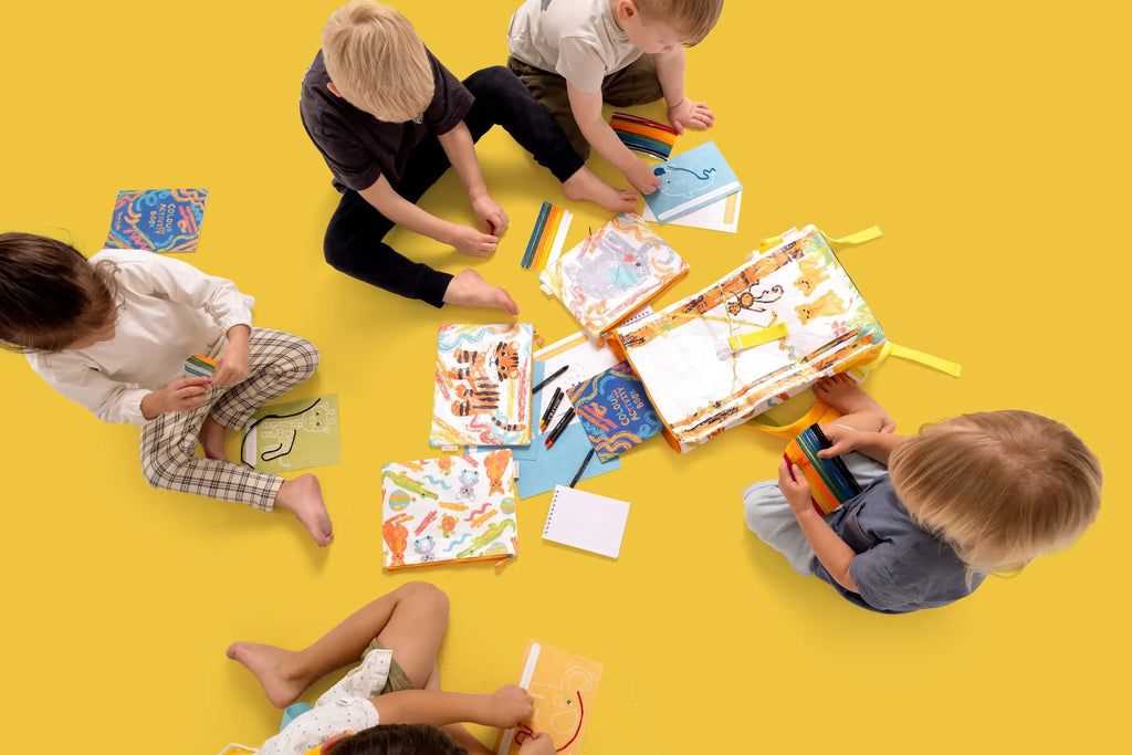 Kids sitting on a yellow floor, playing with colorful wax sticks and drawing on reusable activity books from Two Cubs